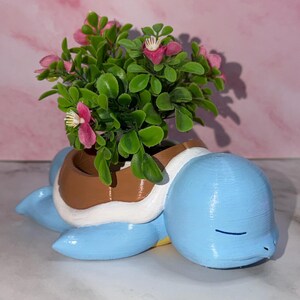 May include: A light blue turtle-shaped planter with a brown and white shell design. The planter contains a small artificial plant with green leaves and pink flowers. The turtle has a smooth, rounded head with closed eyes and a horizontal mouth line.