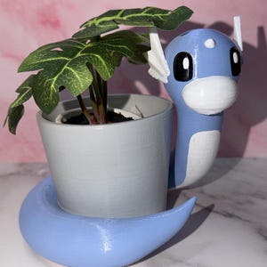 May include: A light blue and white planter shaped like a cartoon dragon, holding a small gray pot with a green leafy plant. The planter has a curved base and white accents. The plant has dark green leaves.