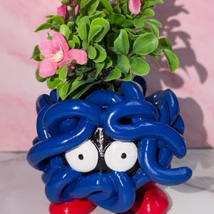 May include: A blue, spherical planter featuring a character design with large white eyes, black facial features, and two red feet. The planter is filled with a small artificial plant that has green leaves and several small pink flowers.