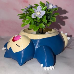 May include: A blue, beige, and white Snorlax planter with a small green plant and purple flowers. The planter is a 3D-printed design of the Pokémon character Snorlax, lying down. The flowers and plant are artificial.