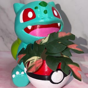 May include: A teal Bulbasaur figurine featuring red eyes, a pink mouth, and white teeth. The character holds a red, white, and black Poké Ball planter containing artificial green and pink variegated leaves. The surface is smooth and painted.