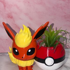 May include: A decorative planter featuring a red and yellow Flareon character figure attached to a round red, white, and black Poké Ball container. A small green air plant is placed inside the spherical opening of the Poké Ball planter.