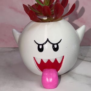May include: A white spherical planter shaped like a ghost character with a wide open mouth, sharp white teeth, and a protruding pink tongue. The planter contains a small succulent with dark red leaves, featuring two small side points.