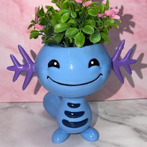 May include: A light blue axolotl-shaped planter with purple external gills, a wide smiling face, and dark blue horizontal stripes on its torso. The planter holds a small artificial plant with green leaves and tiny pink flowers.
