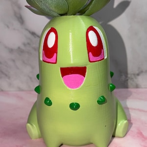 May include: A light green, cylindrical planter shaped like a cartoon creature with a wide, smiling mouth, pink tongue, and large red eyes with white pupils. The body features small, dark green protrusions and holds a green artificial succulent plant.