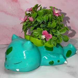 May include: A teal creature-shaped planter with dark green spots, a small white tooth, and closed eyes. A bright green bulb-shaped container sits on its back, filled with artificial green foliage and small pink flowers with white centres.