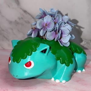 May include: A teal creature-shaped planter with a dark green leafy back, red eyes, and white claws. The planter holds a cluster of light blue and purple artificial flowers, creating a decorative display with a smooth, matte-textured finish.