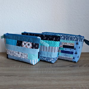 Könnte beinhalten: Drei blaue und weiße gestreifte Reißverschlusstaschen mit nautischem Stoff. Die Taschen sind aus Patchwork-Stoff mit Ankern, Sternen und anderen nautischen Motiven. Die Taschen sind in einer Reihe auf einer Holzoberfläche angeordnet.