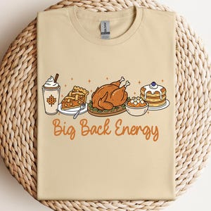 Puede incluir: Camiseta beige con un gráfico de alimentos de Acción de Gracias, que incluye una bebida de café, pastel, pavo, puré de papas y panqueques. El texto "Big Back Energy" está impreso en naranja. La camiseta se muestra sobre un mantel individual circular tejido.