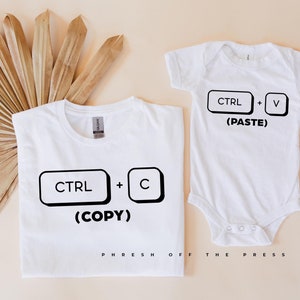 Op de afbeelding: Een wit T-shirt met de tekst "CTRL + C (COPY)" en een witte romper met de tekst "CTRL + V (PASTE)". Beide shirts hebben zwarte toetsen van een toetsenbord.