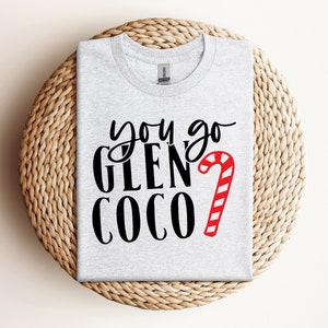 Może przedstawiać: Jasnoszara koszulka z napisem "you go GLEN COCO" w czarnym kolorze i czerwono-białą laską cukrową. Koszulka jest złożona na okrągłej, plecionej podkładce.