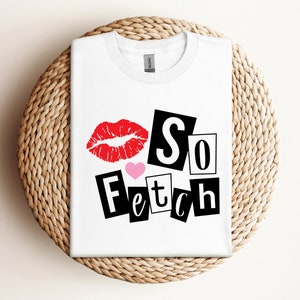 Może przedstawiać: Biała koszulka z napisem "So Fetch" w czarnym, blokowym kroju pisma, nadrukiem czerwonych ust i różowym sercem. Koszulka jest złożona na tkanej, okrągłej powierzchni.