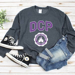 May include: A dark gray sweatshirt with a purple "DCP" logo and a purple and white vintage-style logo with a castle and the text "College Program Est. 1981".