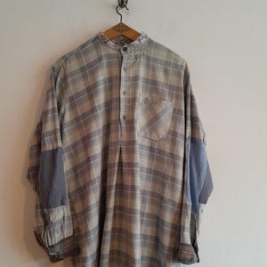 May include: A long-sleeved, button-down shirt featuring a grey and beige plaid pattern. The shirt has a mandarin collar, a single chest pocket, and solid grey elbow patches on both sleeves. It is displayed hanging on a wooden hanger.