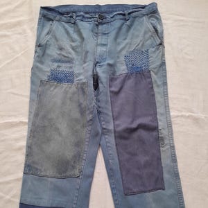 May include: Light blue denim pants featuring a button closure, belt loops, and multiple rectangular fabric patches in varying shades of blue. The garment includes visible sashiko-style stitching details and distressed areas on the front legs.