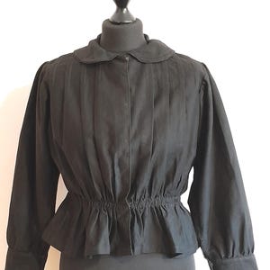May include: Black long-sleeved blouse with a round collar and pleated front. The blouse has a fitted waist with a peplum detail and buttoned cuffs. The garment is displayed on a black mannequin.