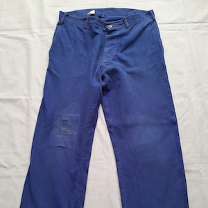 May include: A pair of blue trousers featuring a subtle herringbone pattern, a button and zipper fly closure, belt loops, and a rectangular patch repair on the right leg. The fabric has a visible weave texture throughout the garment.