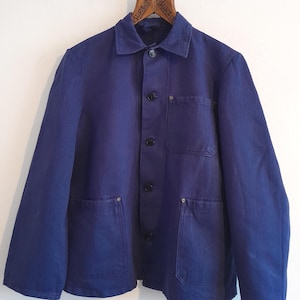 May include: A navy blue work jacket with a button-up front, two front pockets, and a collar. The jacket is hanging on a wooden hanger. The jacket is made of a durable material. The hanger has the word "Burton" on it.