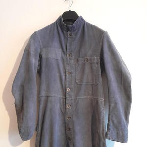 以下が含まれることがあります： スタンドカラーとボタン留めの、くすんだ青色のジャンプスーツ。 ワークウェアスタイルの衣服は、胸に2つのポケットと長袖が特徴です。 ジャンプスーツは、白い壁を背景に黒いハンガーに吊るされています。