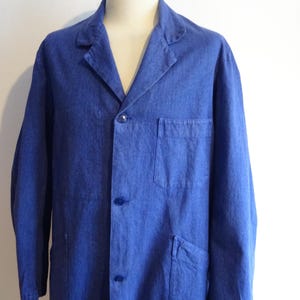 May include: A blue denim work coat with a notched collar, three buttons, and two front pockets. The coat is displayed on a mannequin and has long sleeves. The fabric appears to be a medium weight denim.