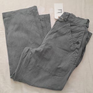 May include: Gray wide-leg trousers featuring a high-waisted design with large front patch pockets, a single button closure, and belt loops. A white rectangular tag attached to the waistband displays the text Emperor of Mercia in black ink.