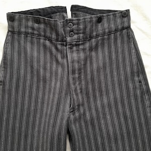 May include: High-waisted trousers in a dark charcoal grey fabric with thin, vertical white pinstripes. The waistband features a double-button front closure and additional buttons along the top edge, with a black interior lining visible.