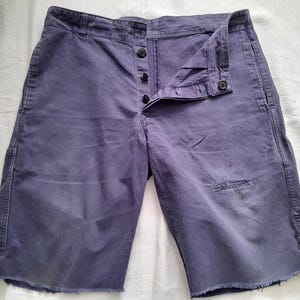 May include: A pair of purple denim shorts featuring a button-fly closure with four visible dark buttons. The shorts have a raw, frayed hemline at the bottom of each leg and a small distressed patch on the front of the right leg near the hem.