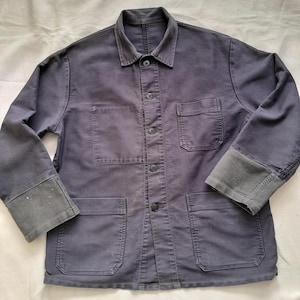 May include: A dark blue work jacket with a button-up front, two chest pockets, and two lower pockets. The cuffs are a darker shade of blue. The jacket has a collar and appears to be made of a durable fabric.
