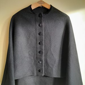 May include: A black cape-style jacket with a round collar and a row of seven dark buttons down the front. The jacket is hanging on a wooden hanger with a decorative metal hook.