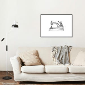 May include: Black and white line drawing of a vintage sewing machine in a black frame. The artwork is displayed on a white wall above a beige sofa with pillows and a patterned throw. A modern floor lamp stands to the left.