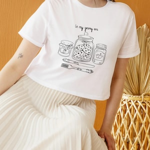 May include: White t-shirt with a black line drawing of three jars, cookies, and a fork and knife. The text "in my granny era" is above the jars. The shirt is a cropped style.