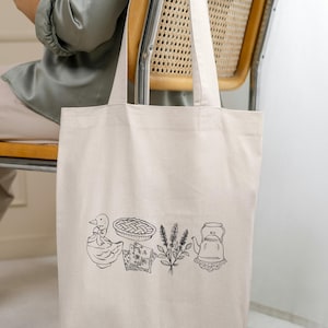 May include: Cream-colored tote bag featuring a black line drawing of a duck, pie, sprigs of lavender, and a teapot. The bag is held by a person sitting in a wooden chair with a woven back. The bag is made of a sturdy canvas material.