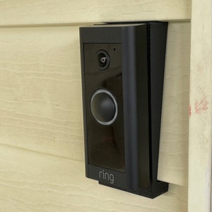 May include: A black Ring doorbell mounted on a light beige siding with the word "ring" in white lettering on the bottom of the doorbell.