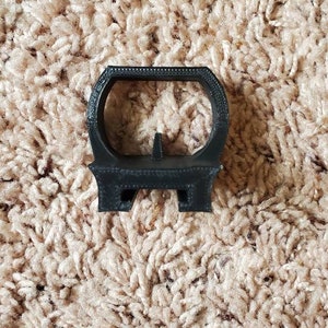 May include: A black plastic ring stand with a small, raised platform. The stand is designed to hold a small object, such as a ring or a bracelet.