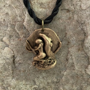 May include: A bronze pendant necklace featuring a sculpted design of two figures embracing. The pendant is attached to a black braided cord.