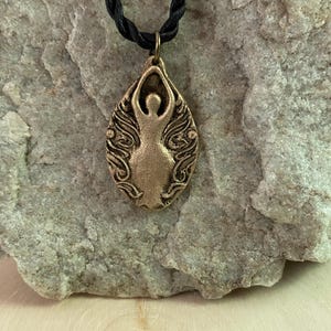 Peut inclure: Un collier avec un pendentif en bronze représentant une figure stylisée dans un cadre ovale. Le pendentif est suspendu à un cordon noir. La figure a les bras levés, entourée de motifs tourbillonnants. Le fond est une pierre texturée.