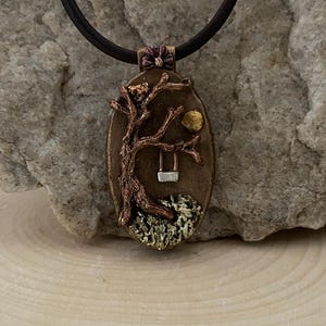 May include: Oval pendant necklace with a bronze-colored scene of a tree, swing, and moon. The pendant has a dark brown leather cord. The tree has a bird perched on a branch. The pendant is set against a stone background.