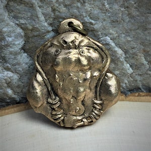 May include: A gold-colored metal pendant depicting a muscular figure with a hooded head. The figure is wearing a rope-like necklace.