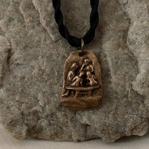 Op de afbeelding: Een bronzen hangerketting met een reliëfsculptuur van een gezin. De hanger hangt aan een zwart koord. De sculptuur toont een gezin van vier personen, zittend op een bank. De hanger is rechthoekig met afgeronde randen.