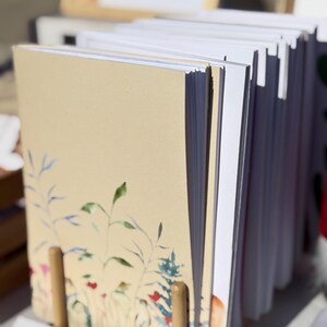 Puede incluir: Una pila de tarjetas en blanco con un diseño floral en la portada. Las tarjetas se mantienen juntas con un soporte de madera.
