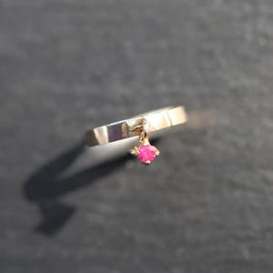 May include: A gold-toned ring featuring a smooth, flat band with a single, small, dangling charm. The charm is a bright pink, faceted gemstone held in a four-prong setting that hangs freely from the top of the ring band.