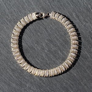 May include: A gold-toned bracelet with a series of oval-shaped links, each encrusted with small, sparkling stones. The bracelet is secured with a clasp. The jewelry is displayed on a dark gray surface.