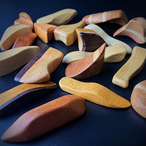 Könnte beinhalten: Eine Sammlung kleiner, glatter, handgeschnitzter Holzobjekte in verschiedenen Formen und natürlichen Holztönen, darunter hellbraune, rötlich-braune und dunkelbraune Nuancen. Jedes Stück hat eine polierte Oberfläche mit deutlicher Maserung.