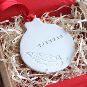 Pode incluir: Um ornamento de cerâmica branca com a palavra "GRANDAD" e um desenho de pena, apresentado numa caixa de presente com papel de seda e fita vermelhos. A caixa é preenchida com palha e está sobre uma superfície tecida.