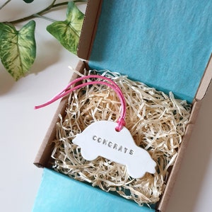 May include: White ceramic car-shaped ornament with the word "Congrats" written on it. The ornament is in a small cardboard box with pink ribbon and shredded paper.