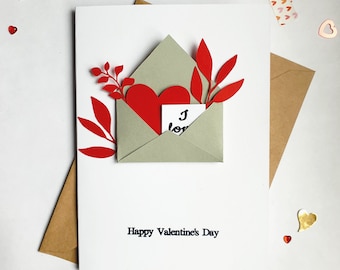 May include: A white card with a red heart and green leaves in a grey envelope. The card says "Happy Valentine's Day" and the envelope has a small white card inside that says "I love you".