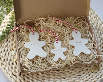 May include: Three white ceramic gingerbread person ornaments with red and white twine hanging from a brown box. The ornaments are personalized with the names "Daddy", "Mummy", and "Aurelia".