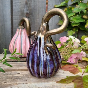 Pode incluir: Duas esculturas de abóbora em cerâmica com corpos canelados e longos caules curvos em bronze metálico. A abóbora da frente apresenta um esmalte roxo e azul profundo, enquanto a abóbora ao fundo tem um acabamento esmaltado rosa e cinza.