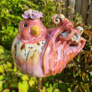 Op de afbeelding: Een keramische vogelsculptuur met een roze en witte body, een gele snavel en een paarse bloemenkroon. De vogel heeft een grillig ontwerp met wervelende details en is gemonteerd op een metalen staaf, perfect voor de tuin.