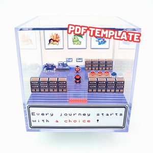 May include: A clear plastic cube display case containing a miniature 3D diorama. The diorama depicts a scene from the Pokemon video game series. The scene includes a Pokemon trainer, a bookshelf, and a sign that reads "Every journey starts with a choice!"
